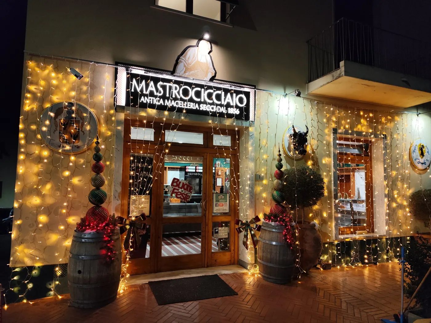 Ingresso di una bottega decorato per Natale con tende di luci sulla facciata, botti, alberi di palline e grandi fiocchi